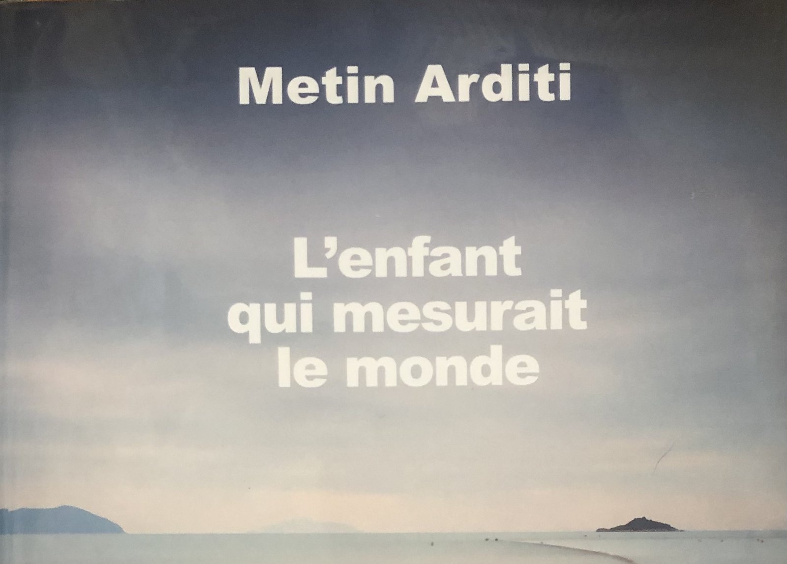 Extrait de la Couverture de L'enfant qui mesurait le monde de Metin Arditi : un filet de pêche court sur une mer tranquille, vers l'horizon, avec deux îles, avec une harmonie de couleurs entre le ciel et la mer.