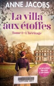 Couverture de La villa aux étoffes, Tome 1, L'héritage, d'Anne Jacobs. Une jeune femme de dos regarde une belle maison au fond de l'allée d'un parc.