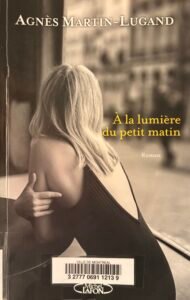 Couverture de À la lumière du petit matin d'Agnès Martin-Lugand : en grand plan, une femme de dos la tête tournée, à la terrasse d'un café