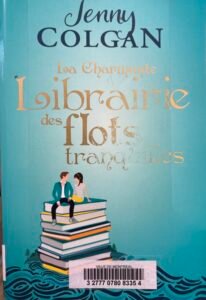Couverture de la charmante libraire des flots tranquilles, de Jenny Colgan : dessin d'un couple assis sur une pile de livres elle-même sur des flots, sur fond bleu turquoise.