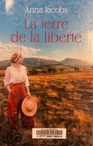 Couverture de La terre de la liberté, par Anna Jacobs ; une femme avec une chemise blanche et un large pantalon, une capeline à la main, regarde le bush australien.